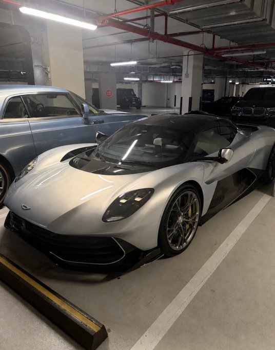 A sleek silver and black hypercar, appearing to be an Aston Martin Valkyrie, parked in an underground garage next to other vehicles.