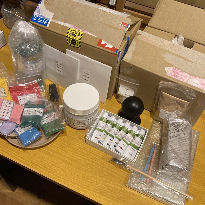 The image shows a wooden table with various art supplies scattered across its surface. The items include paint tubes, brushes, and other craft materials. A black ball is also present among these supplies. In addition to this, there are several boxes on the table that appear to be filled with more art supplies or possibly shipping materials for an artist's project.

The post in question reads: "届いたワクワク 合計約32,000GMT也". This message is likely related to the artist's work and could provide information about the cost of the supplies or a specific art project they are working on. 

In summary, the image showcases an assortment of art materials spread across a wooden table, with some items possibly being part of an artist's project or shipping materials for an upcoming one.