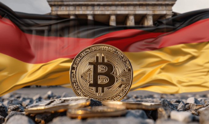 A golden Bitcoin token stands upright on pebbles with a German flag draped behind it and the Brandenburg Gate visible in the background.
