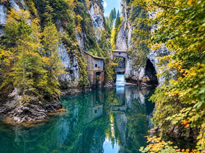 A scenic landscape photograph of a turquoise river gorge flanked by rocky cliffs covered in green and yellow trees, with small wooden structures built into the rocks.