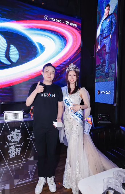 Two people pose together at an event with 'HTX DAO x TRON' branding in the background; the man gives a thumbs-up wearing a TRON shirt, while the woman wears a white gown, crown, and sash.