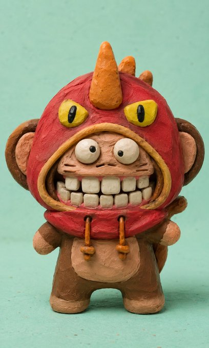 A handcrafted clay figurine of a mischievous monkey wearing a red hood with orange spikes and yellow eyes, displaying a wide toothy grin against a mint green background.
