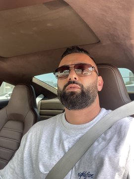 A bearded man wearing sunglasses and a gray "adventure" sweatshirt takes a selfie in a car while wearing his seatbelt, continuing his daily series of selfies as part of "Day 6 of posting a selfie until $HYPE is 3 digits."
