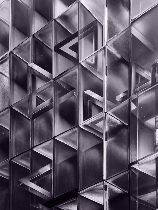 A monochromatic, geometric abstract image of interlocking, metallic three-dimensional triangles and trapezoids in shades of gray and silver.