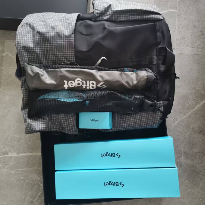 A photograph of an opened black and white grid-patterned bag containing 'Bitget' branded merchandise, including a mesh bag, a small turquoise pouch, and two light blue rectangular boxes stacked below.