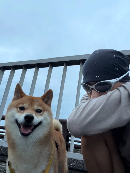 A smiling Shiba Inu dog sits next to a person wearing a black patterned cap and sunglasses, posed together on a balcony or terrace.