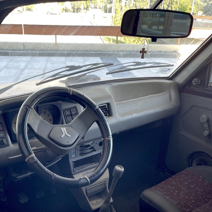 An interior view of an older vehicle showing the dashboard, steering wheel with Renault logo, manual gear shift, rearview mirror with cross hanging, and worn fabric seats.