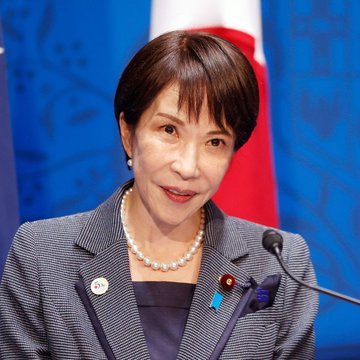 The image features a woman wearing a gray suit jacket and pearl necklace, standing behind a microphone at an event. She appears to be speaking or addressing an audience. The background of the image has a blue color with red and white stripes, which is reminiscent of the flag design seen in the image. The caption reads "Japan Just Officially Accepted Crypto As A MAINSTREAM FINANCIAL ASSET", indicating that Japan has officially recognized cryptocurrency as a mainstream financial asset. This recognition suggests that the country is embracing digital assets like Bitcoin and Ethereum, potentially leading to changes in tax policies for these assets.