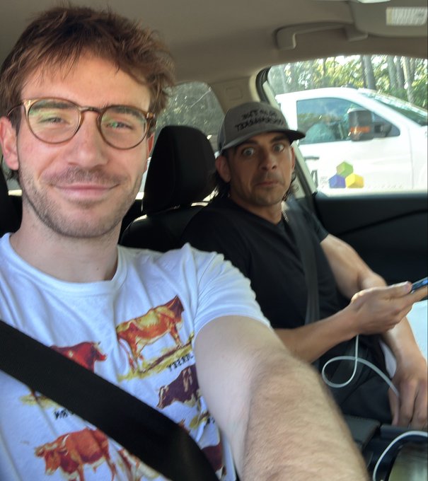 Two men take a selfie inside a car, with the front passenger wearing a cowboy-print t-shirt and glasses while the driver sports a gray cap reading "THEY CALL ME TRASHMAN-91" and looks wide-eyed and excited at the camera. The image's adventurous, road-trip energy matches the post's playful tone of being whisked away to an unexpectedly beautiful mystery location by @CountryBitcoin.