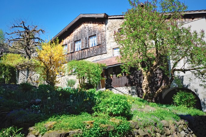 A traditional stone and wood building with wooden shutters sits on a hillside surrounded by lush greenery and trees under a clear blue sky.