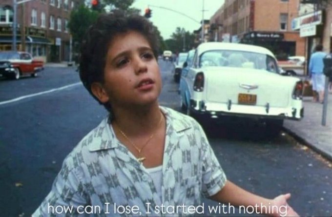 A still from a movie showing a young boy with curly hair wearing a patterned shirt and gold cross necklace on a city street with vintage cars; the subtitle reads "how can I lose, I started with nothing."