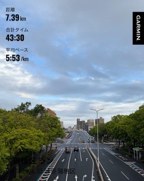 The image shows a city street with cars driving down it, surrounded by trees. The sky above appears cloudy, suggesting an overcast day. In the background of the photo, there are buildings visible, indicating that this scene takes place in an urban area. The main focus is on the road and its surroundings, providing a sense of depth to the image.