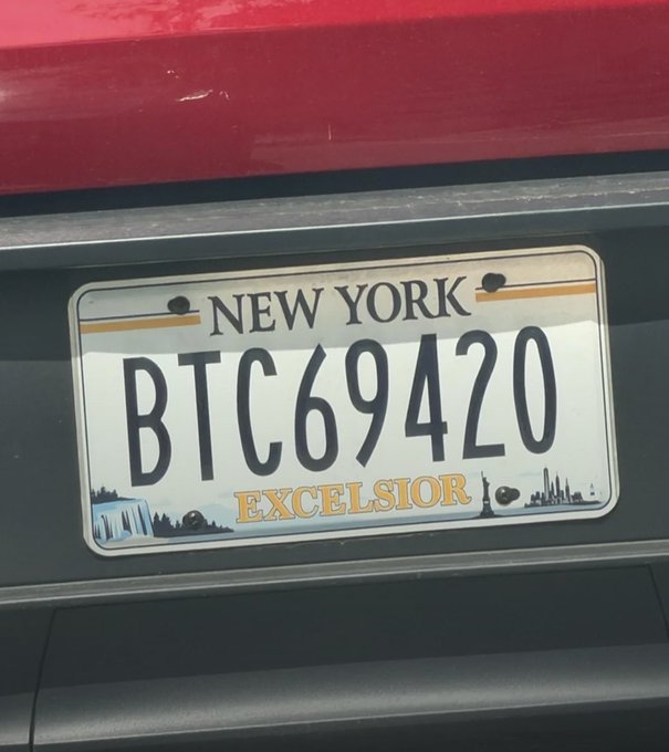 A New York license plate reading 'BTC69420' with the state's 'EXCELSIOR' slogan is mounted on the back of a red vehicle.