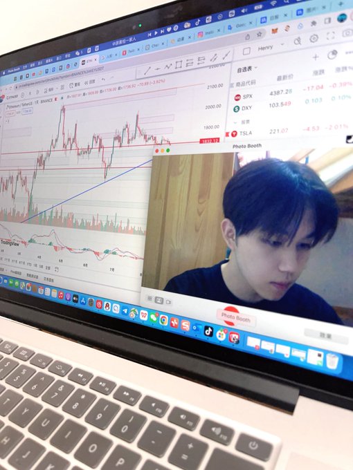 A young man with dark hair sits at a MacBook showing a Photo Booth webcam window overlaid on a stock chart of TSM (Taiwan Semiconductor) trading around 1975, reflecting his self-described identity as a second-level day trader operating from his "HenryKe trading cabin."