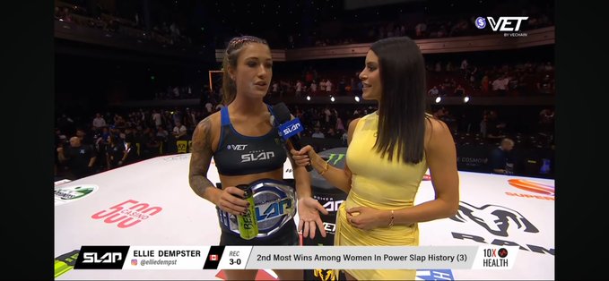 A post-fight interview scene from a Power Slap event shows a female fighter named "ELLIE DEMPSTER" (record 3-0) holding a championship belt while speaking into a SLAP microphone held by a reporter in a yellow dress.