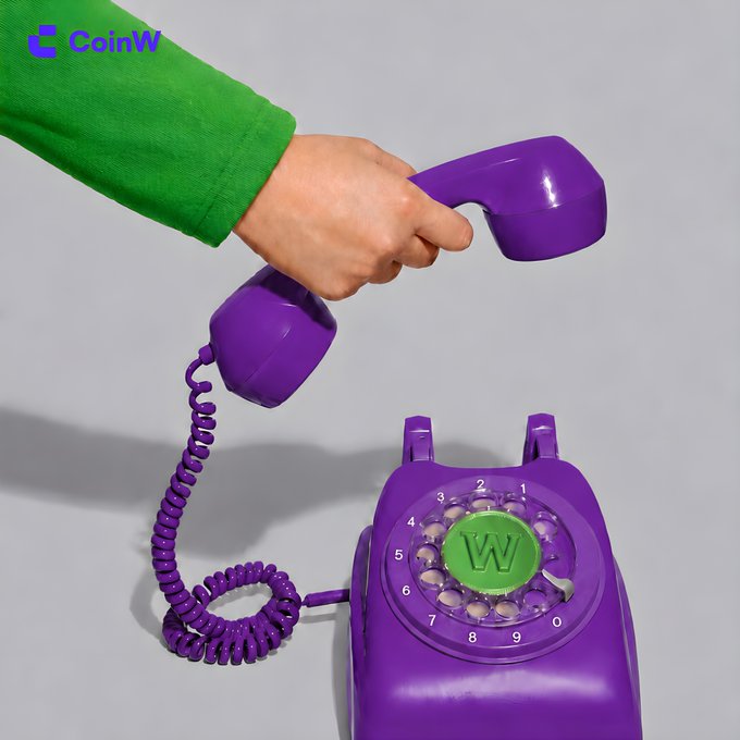 A hand lifts the purple receiver of a rotary phone featuring a green "W" dial center, with the "CoinW" logo visible in the top corner, evoking a vintage communication aesthetic to accompany the casual "GM" (Good Morning) greeting.
