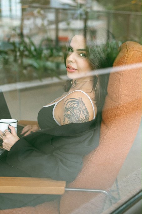 A young woman with dark hair, wearing a black tank top, sits relaxed in an orange chair while holding a mug, showcasing a detailed lion tattoo on her upper arm as she gazes over her shoulder through a window. The image captures a candid, reflective moment that complements the personal and professional tone of her bio, suggesting a thoughtful, well-rounded individual balancing her career in Web3 management with her personal interests like her cats and hobbies.