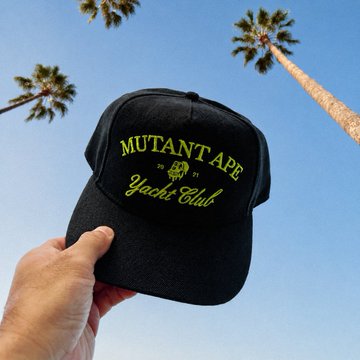 A hand holds up a black baseball cap with neon green embroidery reading "MUTANT TAPE Yachi Club" against a clear blue sky framed by tall palm trees.