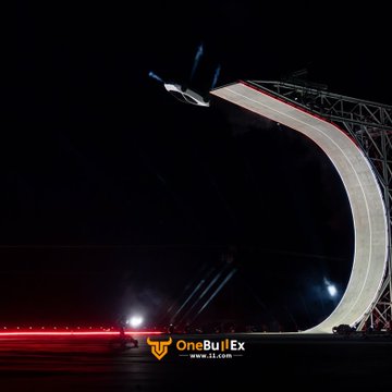A dramatic nighttime photograph captures a vehicle launching off a steep illuminated ski jump ramp, with the 'OneBullEx' logo visible at the bottom.