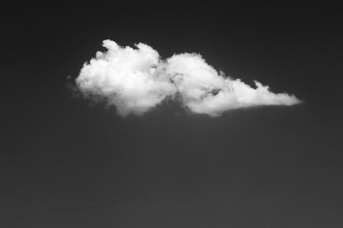 A minimalist black and white photograph showing a single fluffy white cloud isolated against a dark gray sky.