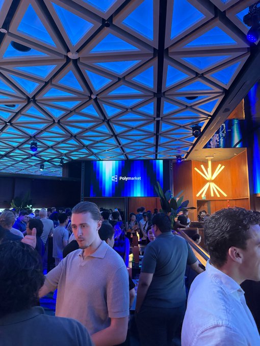 A crowded event in Singapore with a distinctive geometric glass ceiling illuminated in blue, featuring a "Polymarket" display screen in the background. Attendees mingle in what appears to be a modern networking venue, capturing the vibrant atmosphere of a tech or crypto gathering in the city.