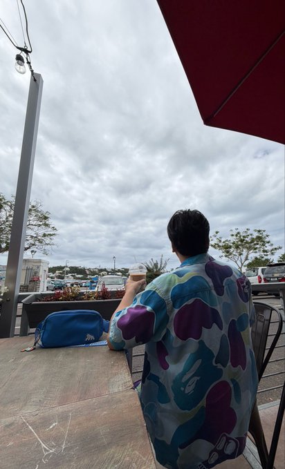 The image shows a person wearing a blue shirt with a camouflage pattern, standing outside at an outdoor cafe or restaurant. The person has their back turned towards the camera, looking up at something above them. There are two umbrellas visible in the scene - one is red and the other is white. A chair can also be seen near the person's feet. In the background, there are several boats of various sizes docked or floating on a body of water. The image includes text that reads "Gm Happy Mutant Monday everyone from Bermuda Repping BAYC on this amazing Island".