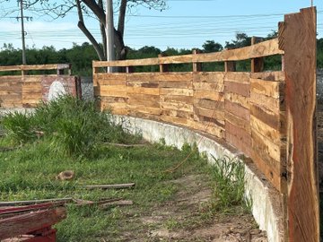 A wooden fence constructed from pallet wood is being built along a concrete wall in a grassy outdoor area.
