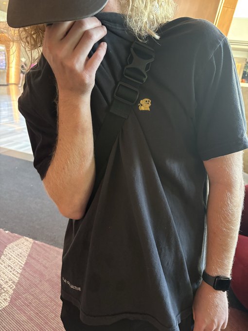 A person with long blonde hair wearing a black t-shirt with 'LA COLLECTIVE' text, a black cap, and a La Collective crossbody strap, standing indoors on a red patterned rug.