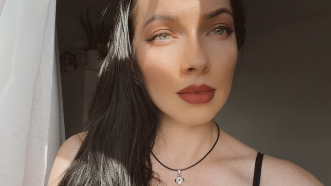 A close-up portrait of a woman with long dark hair, red lipstick, and a black cord necklace with a star pendant, looking directly at the camera in soft lighting.