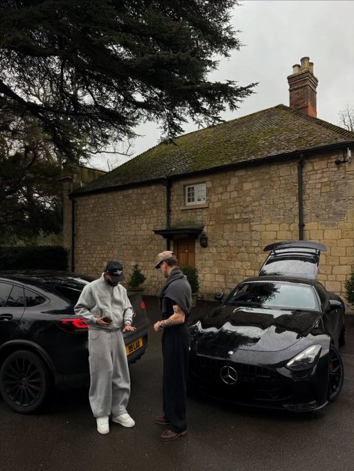 Two men stand beside two black luxury cars (a BMW and a Mercedes) parked in front of a traditional stone house with a moss-covered roof.