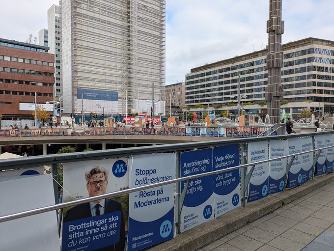A pedestrian bridge in Stockholm displays Moderaterna (Moderate Party) political campaign posters in Swedish, featuring a candidate with glasses and slogans like 'Stoppa bolöneskatten' and 'Ansträngning ska löna sig.'