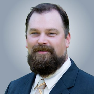 A professional headshot of a bearded man in a dark suit and tie smiling against a plain grey background.