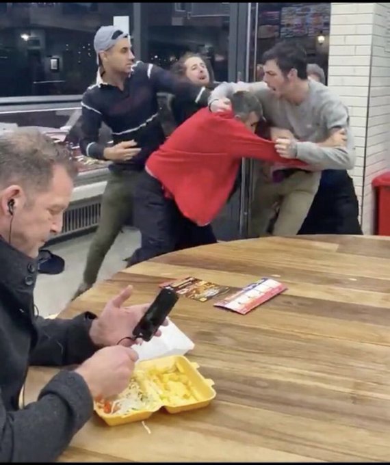 A humorous image of people mock-fighting in the background of a fast-food restaurant while a man in the foreground calmly eats fries from a styrofoam container and looks at his phone.
