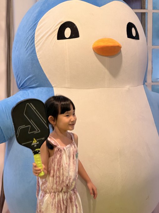 A young girl holding a black pickleball paddle stands in front of a large blue and white penguin mascot plush character.