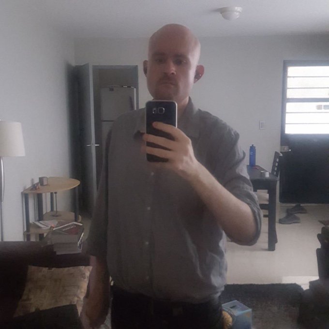 A bald man in a gray button-up shirt takes a mirror selfie in a sparsely furnished apartment, capturing himself on a couch with a refrigerator and living room visible behind him. The image reflects the vulnerability and isolation expressed in the caption, where he lists his struggles—being single, unemployed, and losing six figures in memecoin trading—at 43 years old.