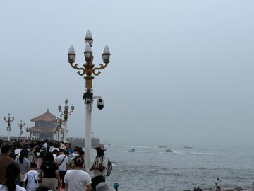 Tourists crowd the famous Zhanqiao Pier in Qingdao on a foggy day, with the iconic ornate lamppost in the foreground and the Huilan Pavilion barely visible through the thick mist in the distance.