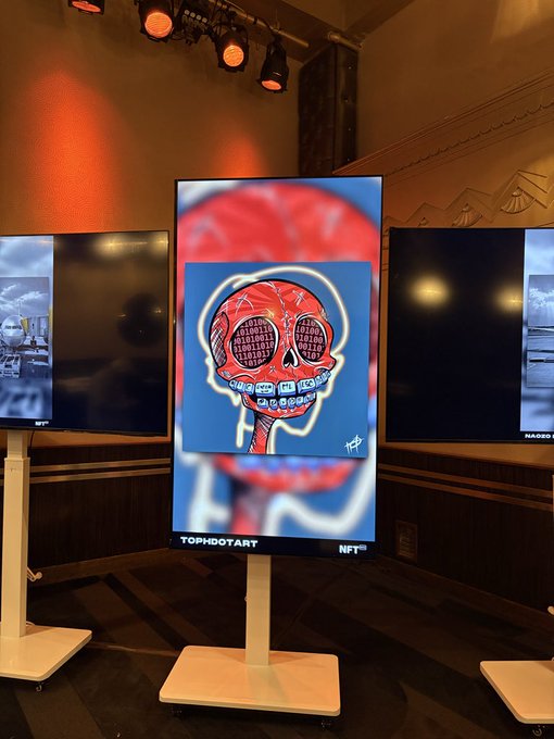 A digital display screen in an art exhibition shows a neon skull artwork with text including binary code in the eyes, displayed on a stand labeled 'TOPHOOTART' and 'NFT'.
