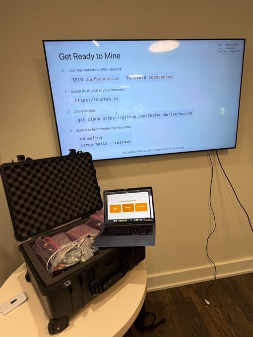 A workshop setup with a TV displaying instructions titled 'Get Ready to Mine' including steps to join SSID '256foundation', install Rust, clone Mujina from github.com/256foundation/mujina, and build it with cargo. A laptop sits on an open equipment case filled with electronics.