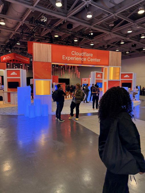 An indoor trade-show floor centers on an orange entrance labeled “Cloudflare Experience Center,” with Cloudflare-branded booths, visitors, and overhead exhibition lighting.