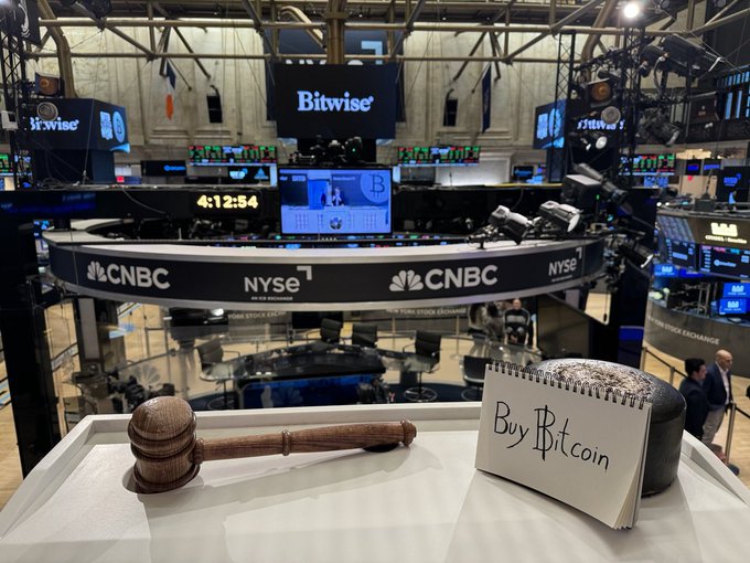NYSE trading floor with CNBC and Bitwise signage visible, featuring a wooden gavel and a handwritten notepad reading 'Buy Bitcoin' in the foreground.