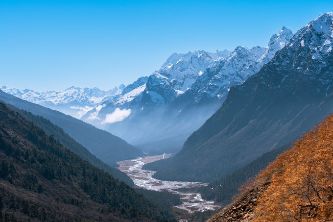 A scenic mountain landscape featuring snow-capped peaks under a clear blue sky, with a winding river valley and forested hillsides, accented by autumn-colored foliage in the foreground.