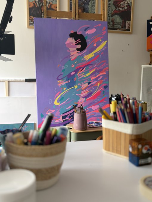 An artist's studio featuring a colorful abstract painting on an easel depicting a silhouette with swirling vibrant colors against a purple background, with baskets of colored pencils in the foreground.