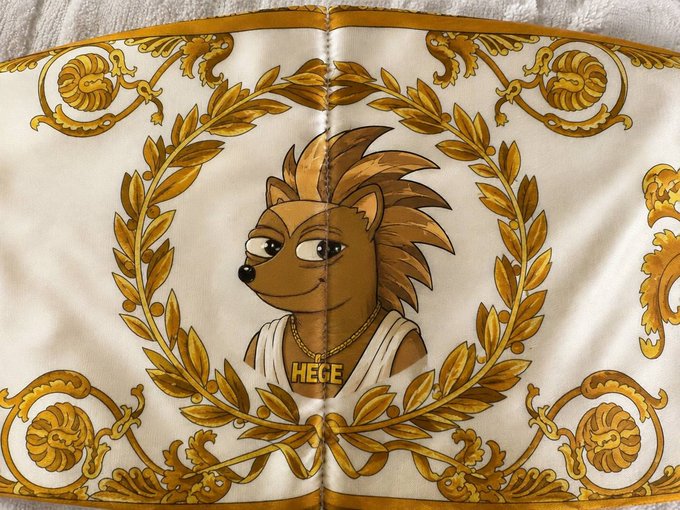 A cream and gold patterned fabric features a hedgehog character wearing a gold chain necklace with the name 'HEGE' on its pendant.