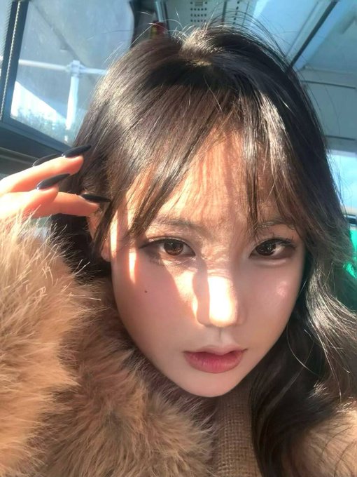 A close-up selfie of a young woman with dark hair, bangs, and pink lipstick, wearing a fluffy brown coat and long black nails, bathed in warm sunlight.