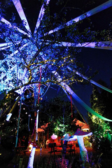 Vibrantly illuminated trees draped in blue, white, and purple lights set a magical nighttime scene perfect for saying "Gn folks" as the evening winds down. Colorful ribbons, lanterns, and glowing decorations hang from the branches, creating a whimsical outdoor gathering space.