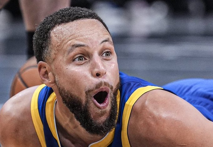 A close-up photograph of basketball player Stephen Curry in a Golden State Warriors jersey, showing a shocked expression with his mouth open and eyes wide.