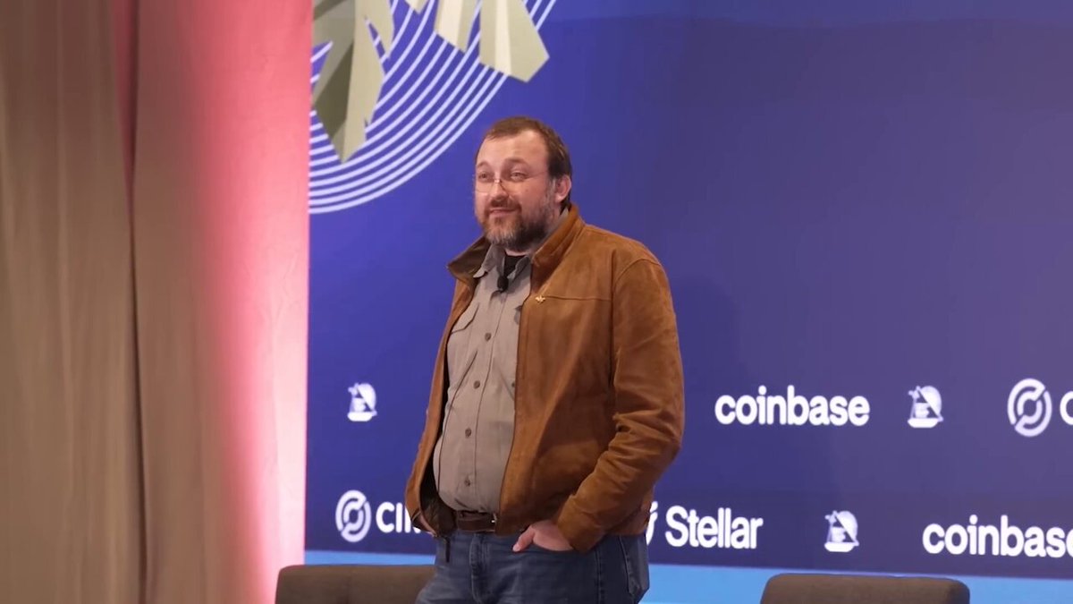 The image features a man standing next to a blue wall with a sign that says "coinbase" on it. The man appears to be wearing a brown jacket, and he has his hands in his pockets while looking at the camera. In the background, there is another person who seems to be attending an event or gathering related to cryptocurrency.