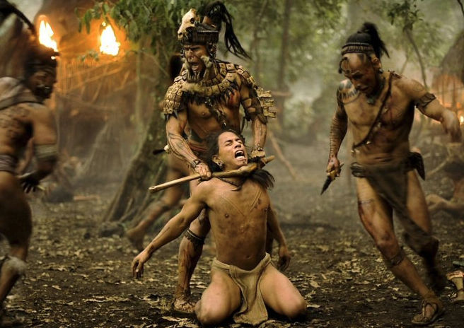 Cinematic still depicting a tribal warrior scene with armored indigenous fighters capturing a kneeling man in a jungle setting with fire in the background.
