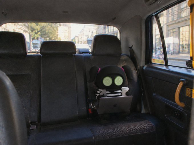 A black stuffed animal with large green button eyes sits in the back seat of a taxi or vehicle, working on an Apple MacBook laptop alongside a coffee cup.
