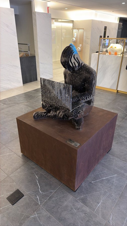 A metal sculpture of a seated figure wearing a blue hat and looking at a laptop, displayed on a brown pedestal in a modern interior with marble flooring.